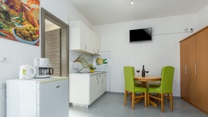 Een compacte keuken met witte kasten, een kleine koelkast, een koffiezetapparaat en een eethoek met twee groene stoelen en een tafeltje.
