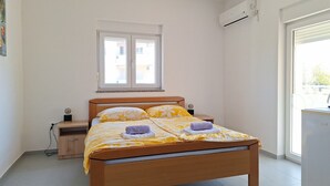 Een enkel bed met houten frame, een nachtkastje met lamp, een raam met zonwering en een airconditioning op de muur.