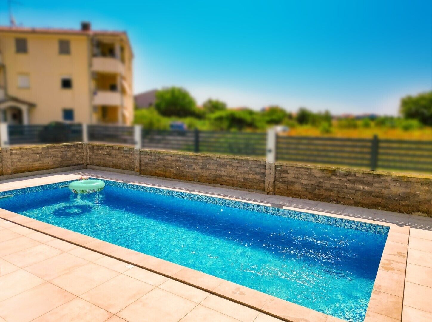 A rectangular swimming pool with a tiled deck, a floating ring, and a stone wall.
