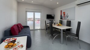 A modern living room with a sofa, dining table, and chairs. There is a TV mounted on the wall and a window with a view.
