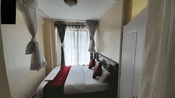 Habitación de hotel estrecha con una cama individual, cortinas blancas y una ventana con cortinas traslúcidas.