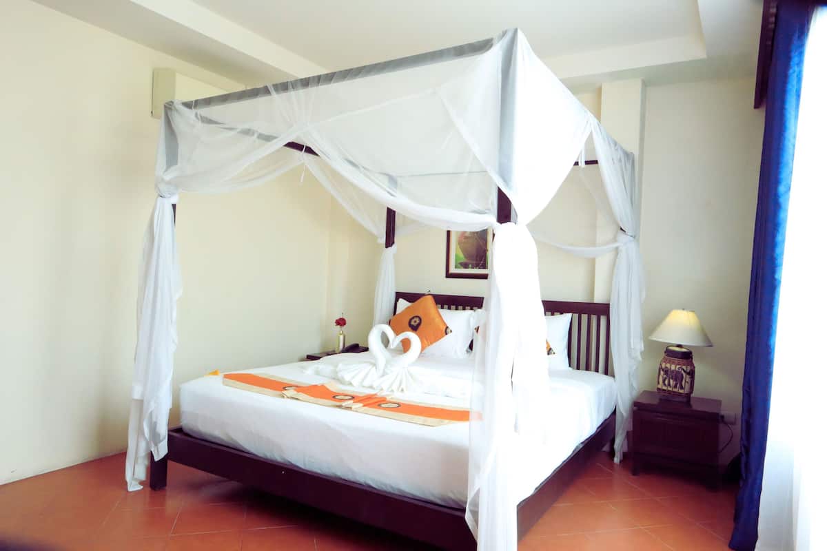 A four-poster canopy bed with a white bedspread and orange decorative elements, a nightstand with a lamp, and a framed picture on the wall.