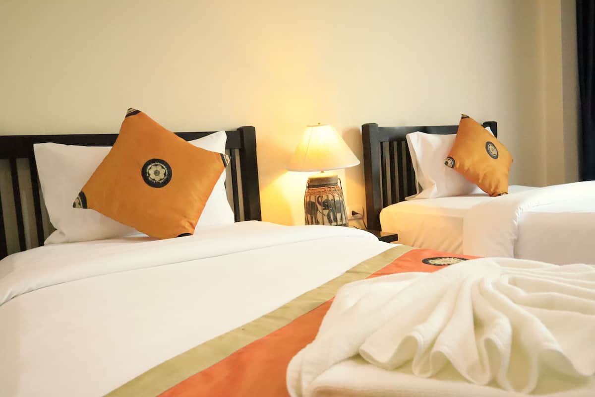 A hotel room with two beds, each with white bedding and orange pillows. There is a small table with a lamp between the beds.