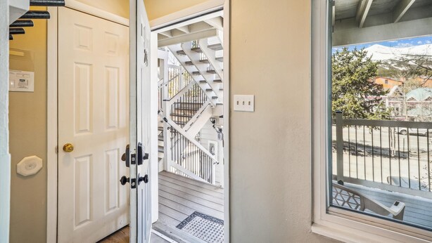 A white door with a brass doorknob, a white wall with a light switch, and a view of a balcony with a metal railing and a building in the background. - Dog-Friendly for Summer|Updated|On S. Main Street (Breckenridge)