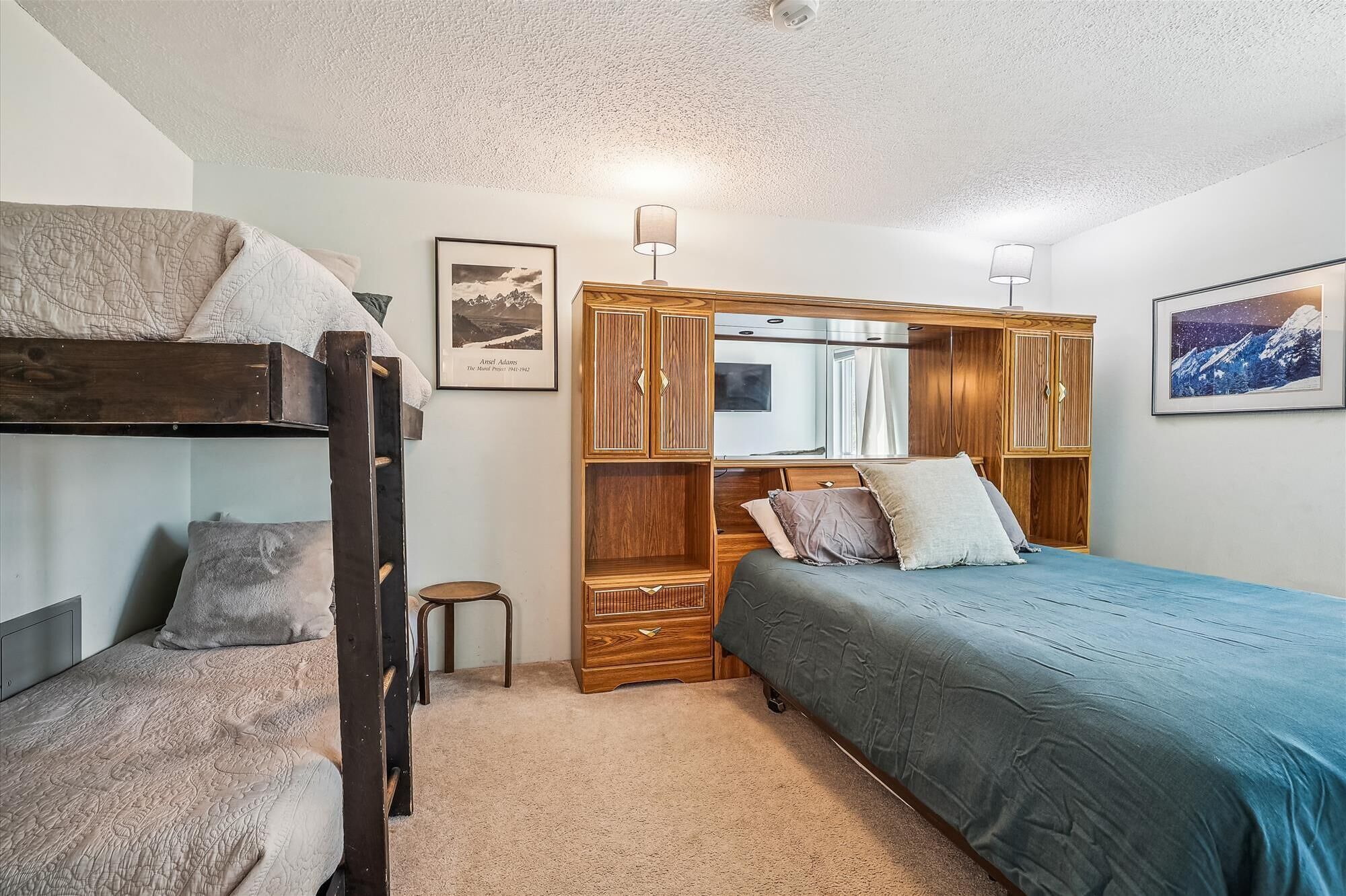 A bedroom with bunk beds, a wooden wardrobe, a small stool, and two framed pictures on the wall.
