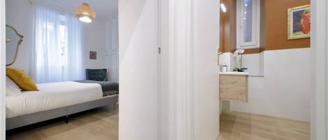 Un dormitorio con cama, mesita de noche, espejo y ventana con cortinas.
