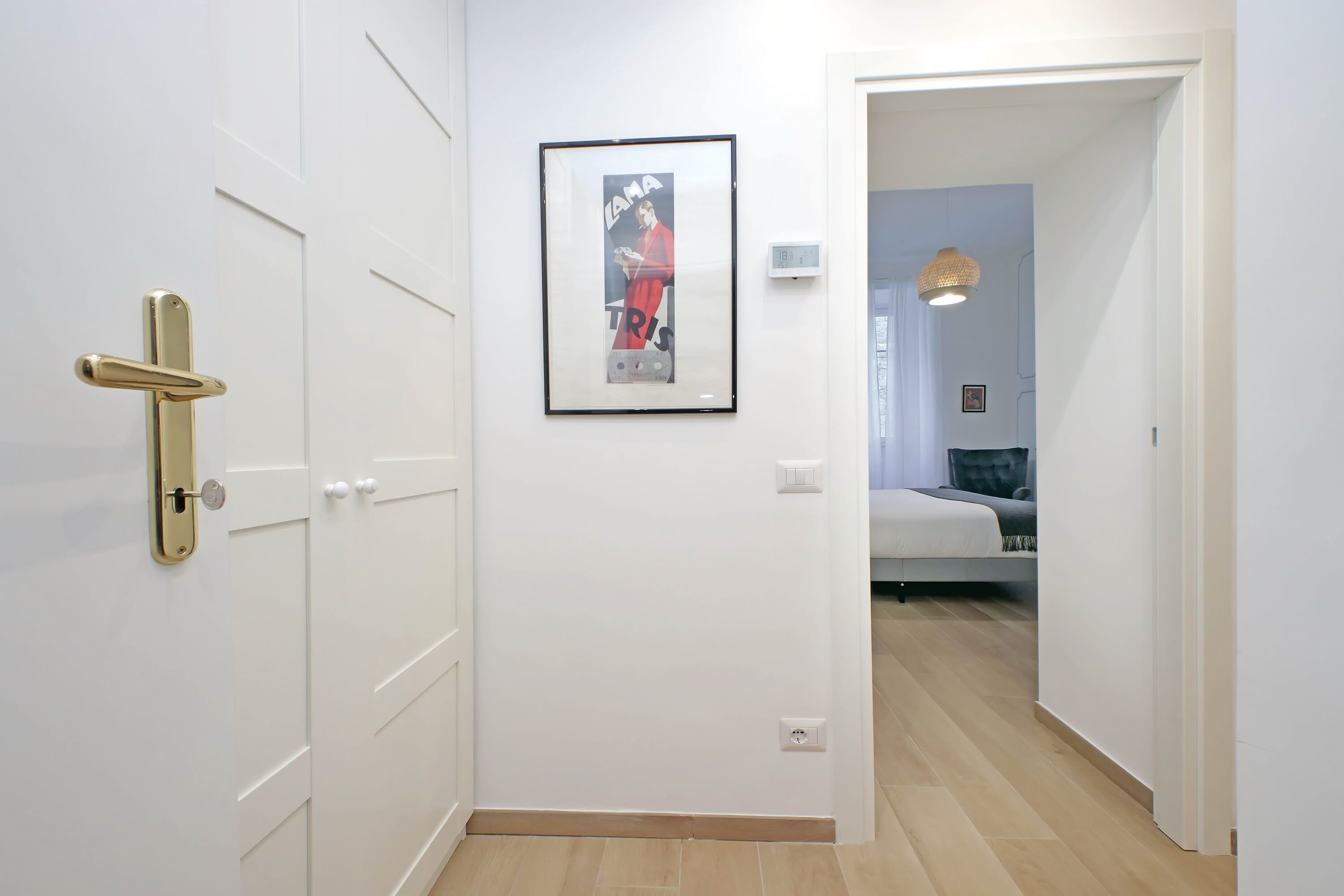 A hallway with a white door, a framed picture on the wall, and a bed in the adjacent room.
