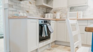 A compact kitchen with white cabinetry, a built-in oven, and a microwave. - The Cow Shed at Pencraig (Holyhead)