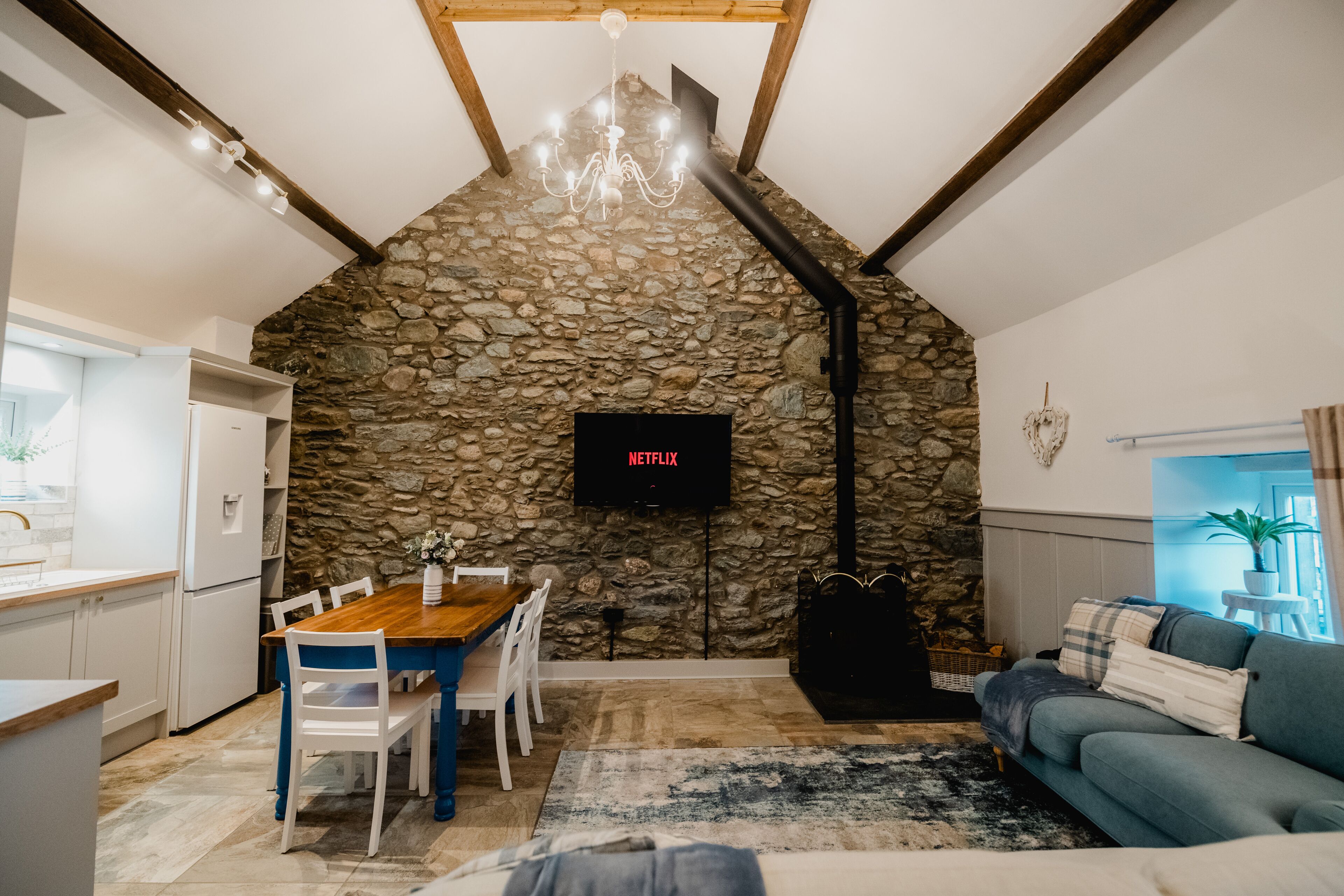 A cozy living space with a stone wall, a wooden dining table, a sofa, and a TV mounted on the wall.
