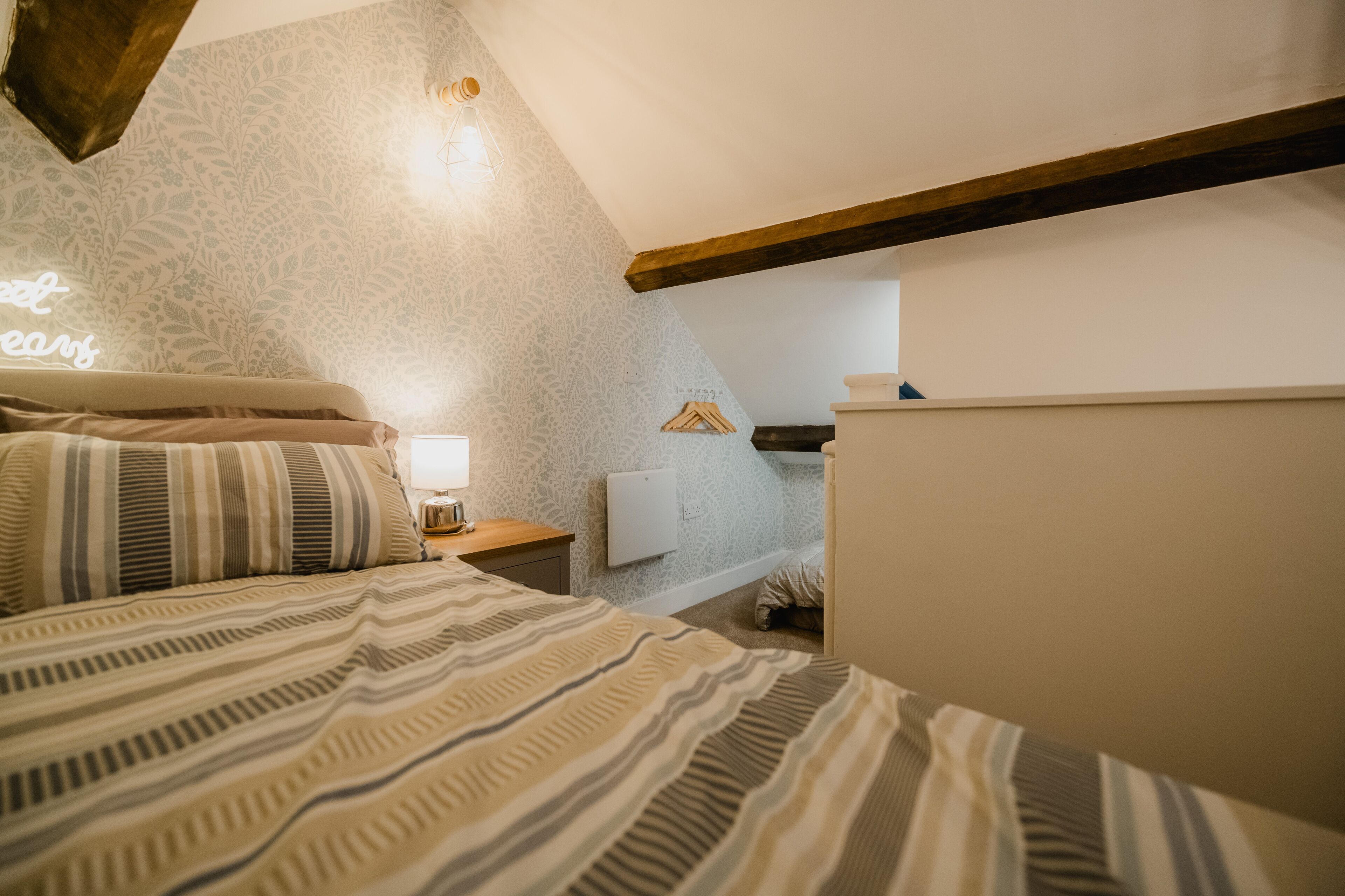 A bedroom with a sloped ceiling, a bed with striped bedding, a bedside table with a lamp, and a wall-mounted light fixture.