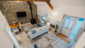 A spacious living area with a stone wall, a sofa, a TV, and a wooden table. - The Cow Shed at Pencraig (Holyhead)