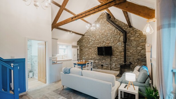 A spacious living area with a stone wall, a sofa, a dining table, and a staircase. - The Cow Shed at Pencraig (Holyhead)