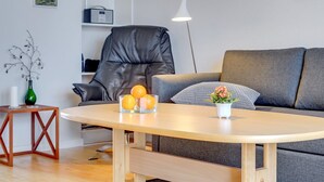 Un soggiorno moderno con una poltrona in pelle, un divano grigio, un tavolino in legno con frutta e fiori, un piccolo tavolino e una libreria con oggetti decorativi.