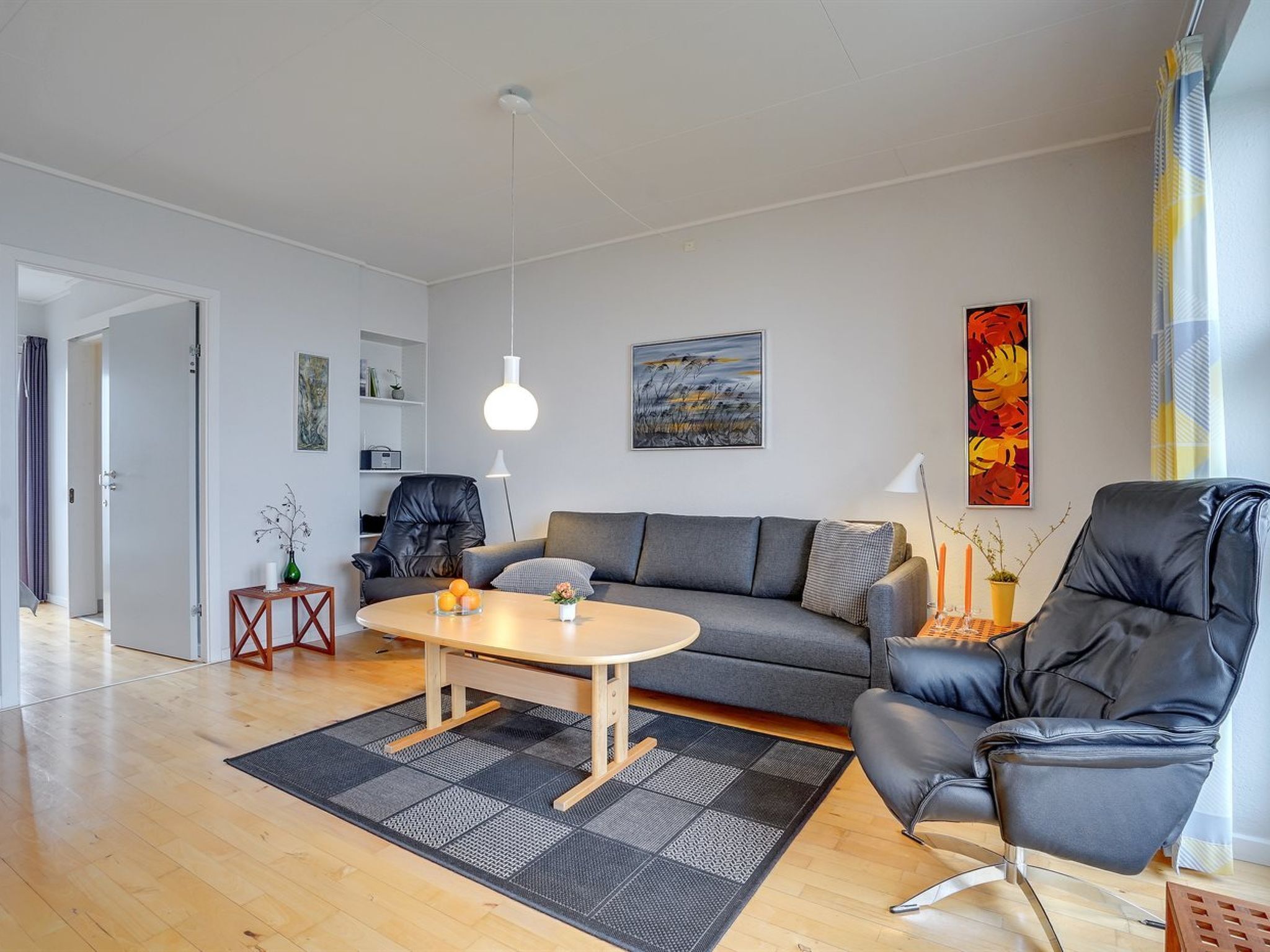 A modern living room with a grey sofa, a black leather chair, a wooden coffee table, and a black and grey rug. There is a painting on the wall and a window with curtains.