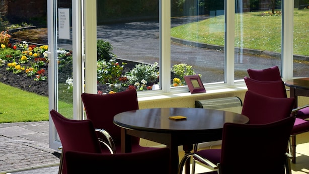 A conservatory with a glass door, a round table, and red chairs. - The Salfordian Hotel (Southport)