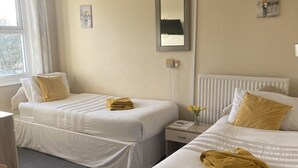 A hotel room with two beds, a bedside table, a chair, a window with curtains, and a wall-mounted mirror. - The Salfordian Hotel (Southport)