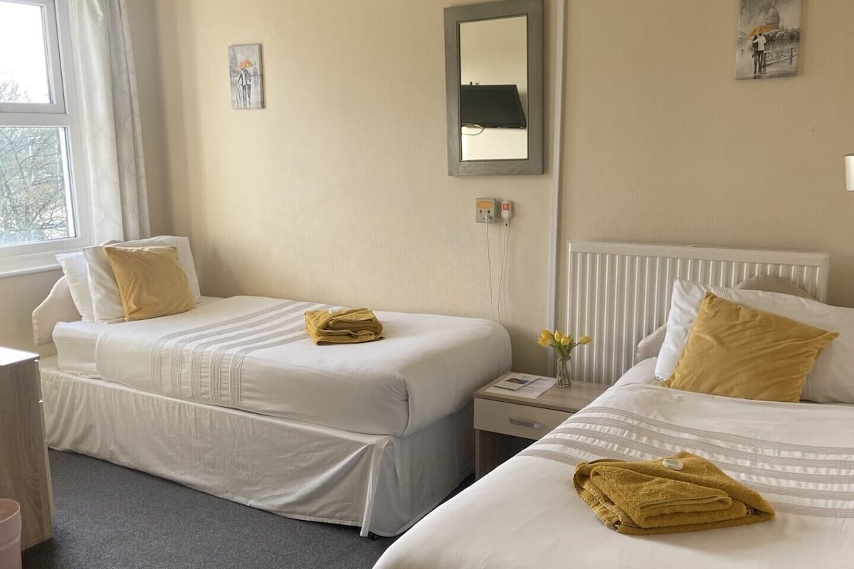 Ein Hotelzimmer mit zwei Betten, einem Nachttisch, einem Sessel, einem Fenster mit Vorhängen und einem wandmontierten Spiegel.