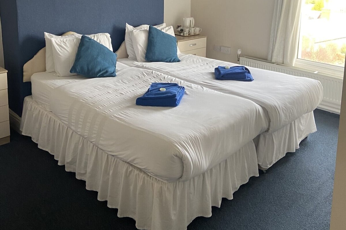 Ein Doppelbett mit weißer und blauer Bettwäsche, ein Nachttisch mit Lampe und ein Fenster mit Vorhängen.