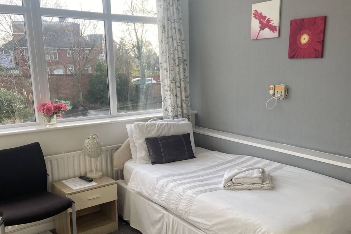 Ein Schlafzimmer mit Bett, Stuhl, Nachttisch, Lampe und einem Fenster mit Vorhängen.
