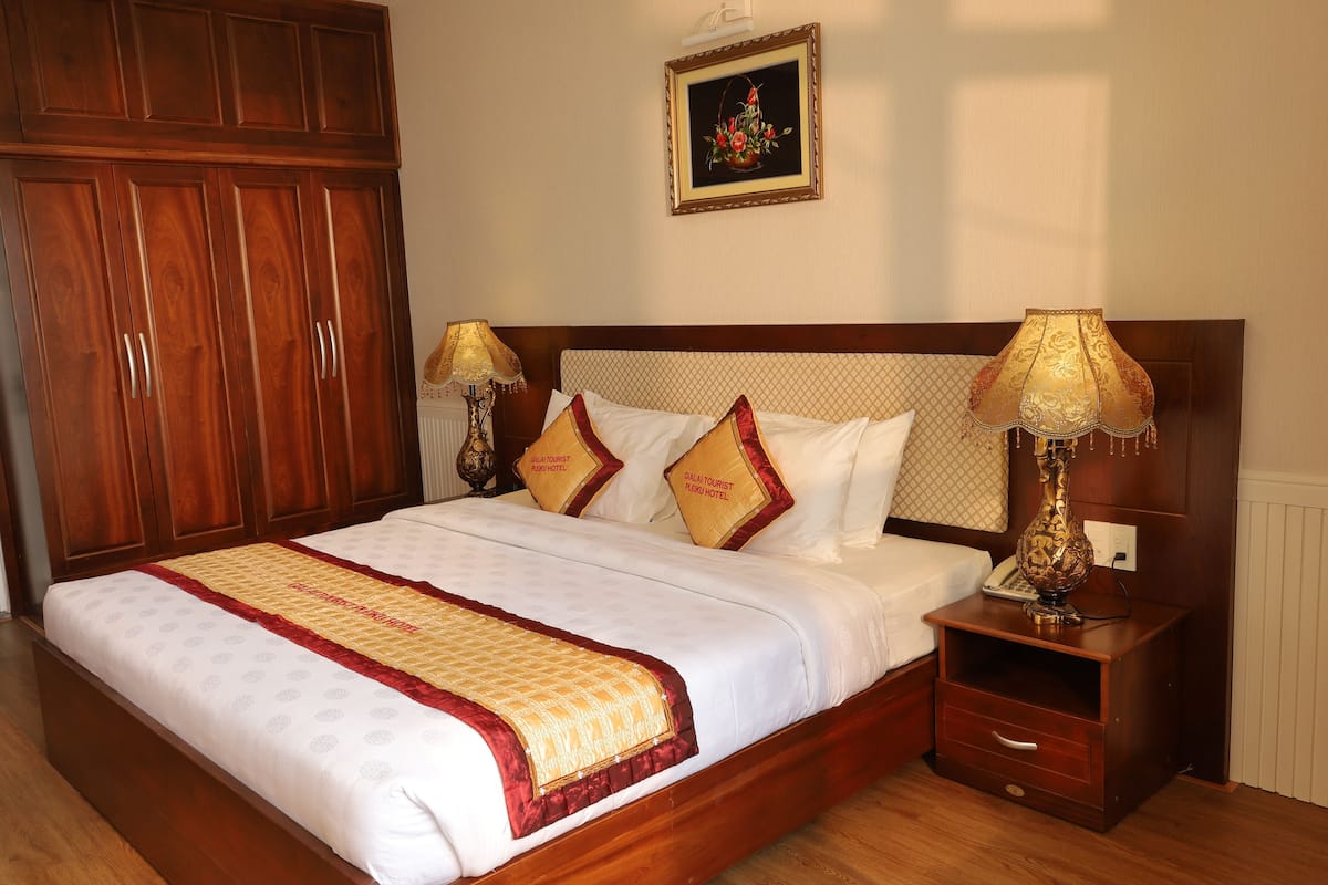A neatly made bed with a white and gold bedspread, two matching pillows, a wooden nightstand with a lamp, and a large wardrobe.