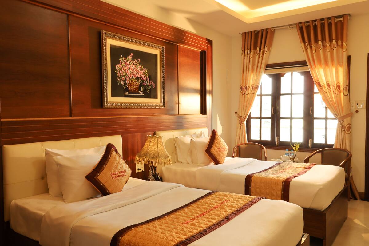 A hotel room with two beds, a wooden headboard, a framed floral painting, and a window with curtains.