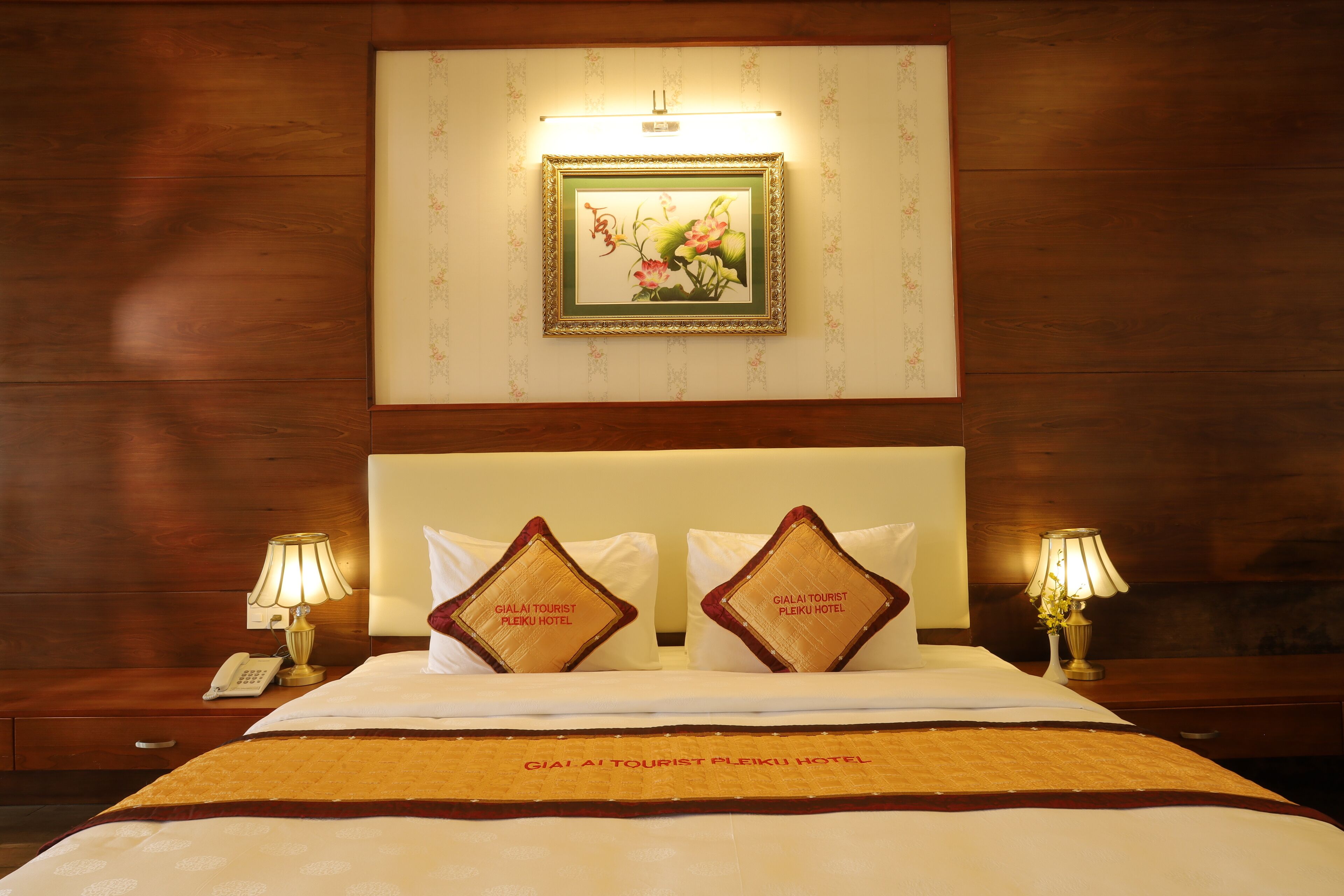 A hotel room with a neatly made bed, two pillows with the hotel name, bedside lamps, a nightstand with a phone, and a framed picture on the wall.