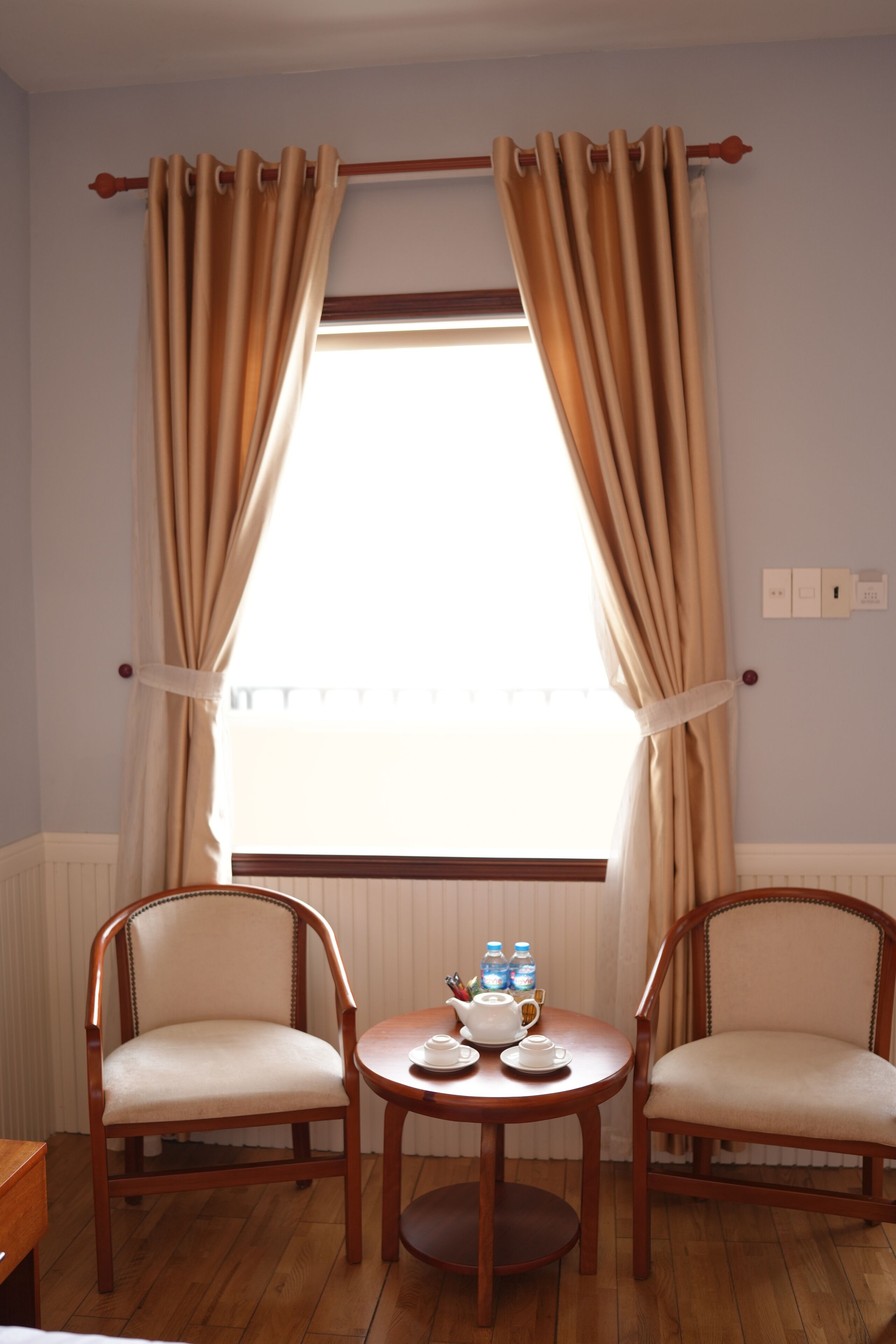 Una habitación con dos sillas de madera, una mesa redonda pequeña con tetera y tazas, y una ventana con cortinas color beige.