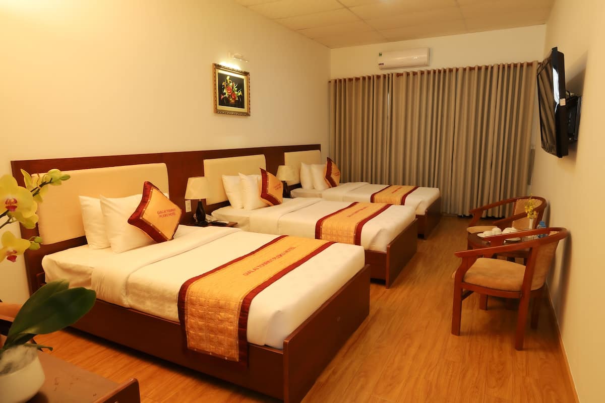 A hotel room with three beds, a TV, a desk with a chair, and a potted plant.