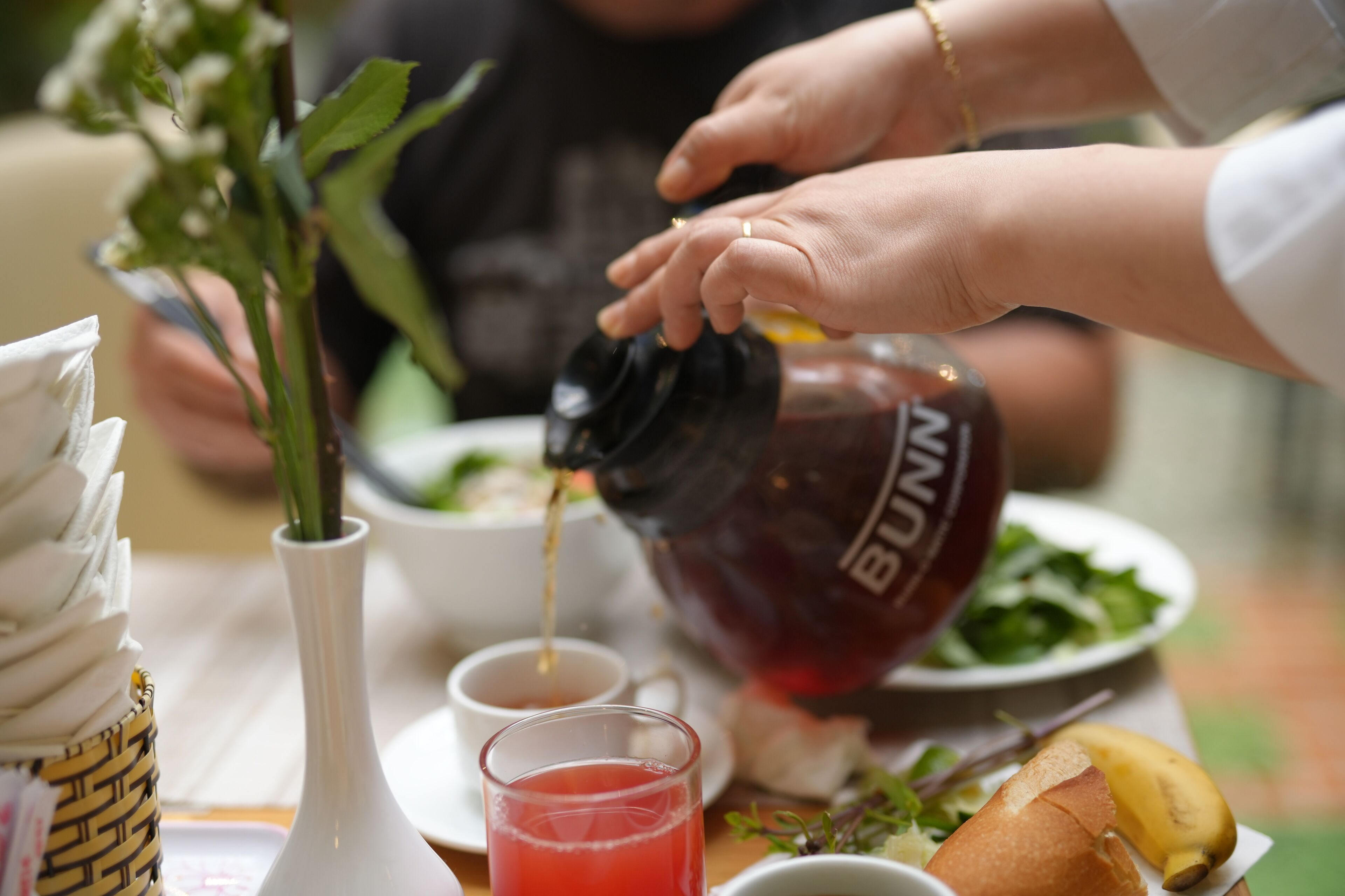 A dining table with a white vase, a glass of red drink, a bowl of salad, and a banana.