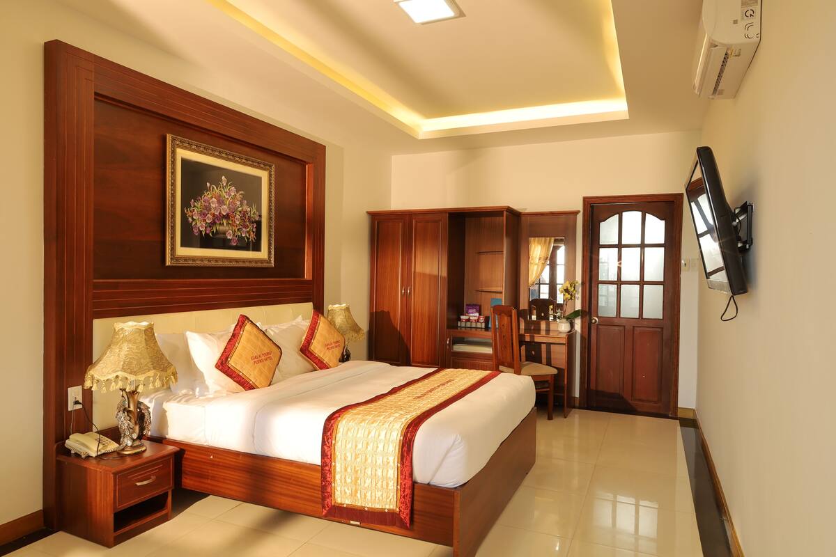 A hotel room with a large bed, wooden headboard, bedside table with lamp, wardrobe, desk, chair, and a television mounted on the wall.