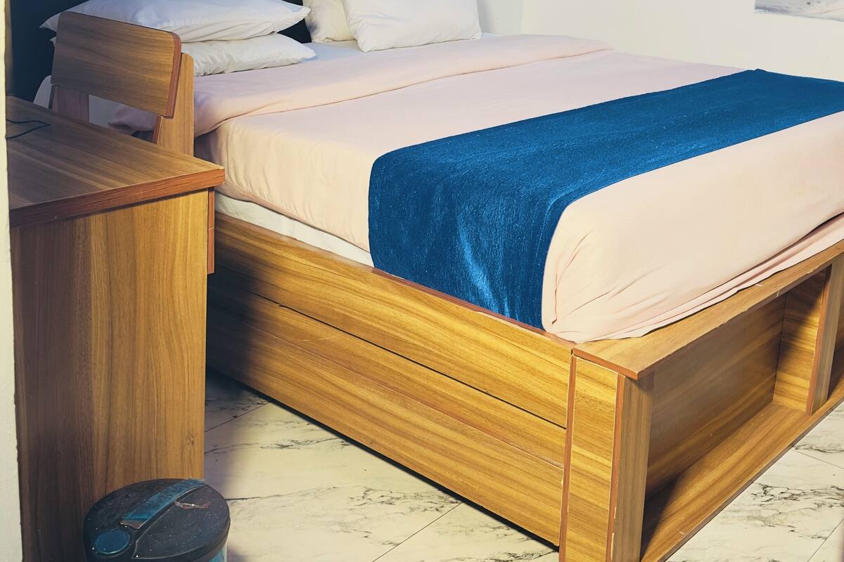 Une chambre avec un lit en bois, un bureau, une poubelle et un rideau.