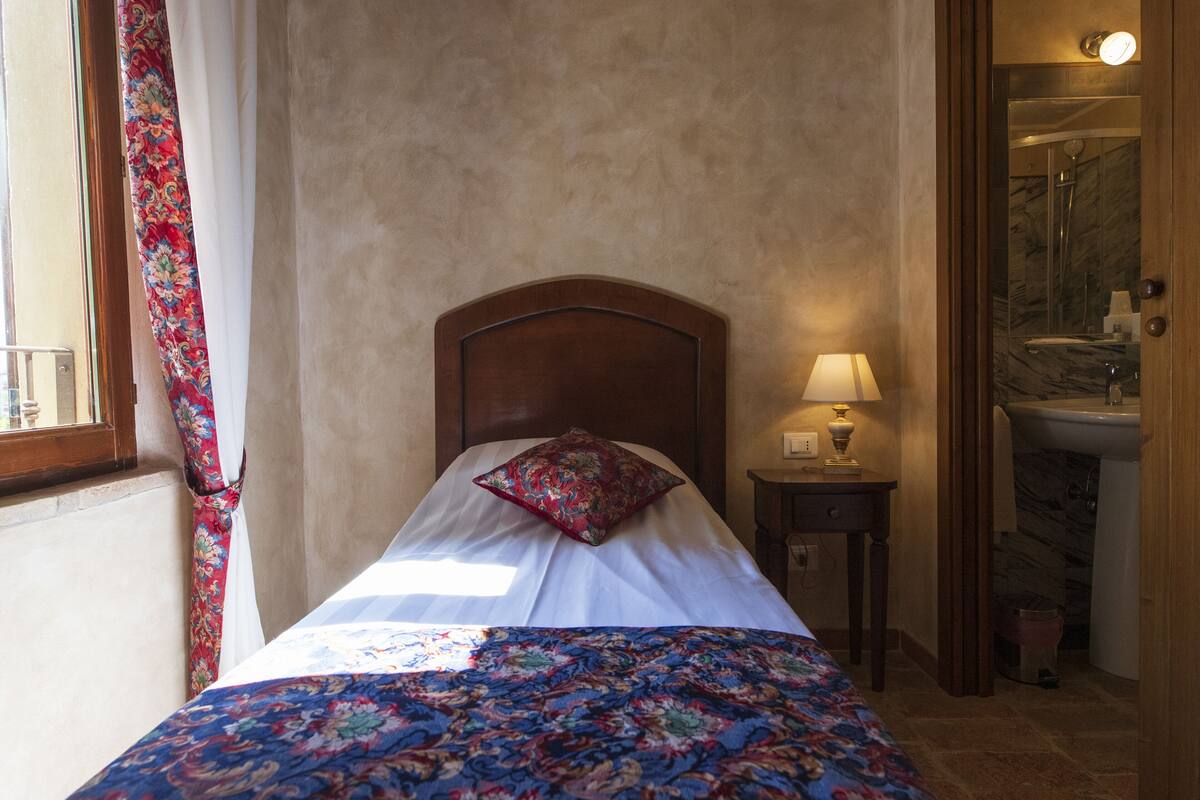 A bedroom with a bed, a nightstand with a lamp, a window with curtains, and a bathroom visible through an open door.