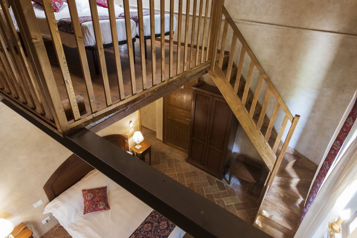 A high-ceilinged room with a wooden staircase, a bed with a floral pillow, and a small table with a lamp.