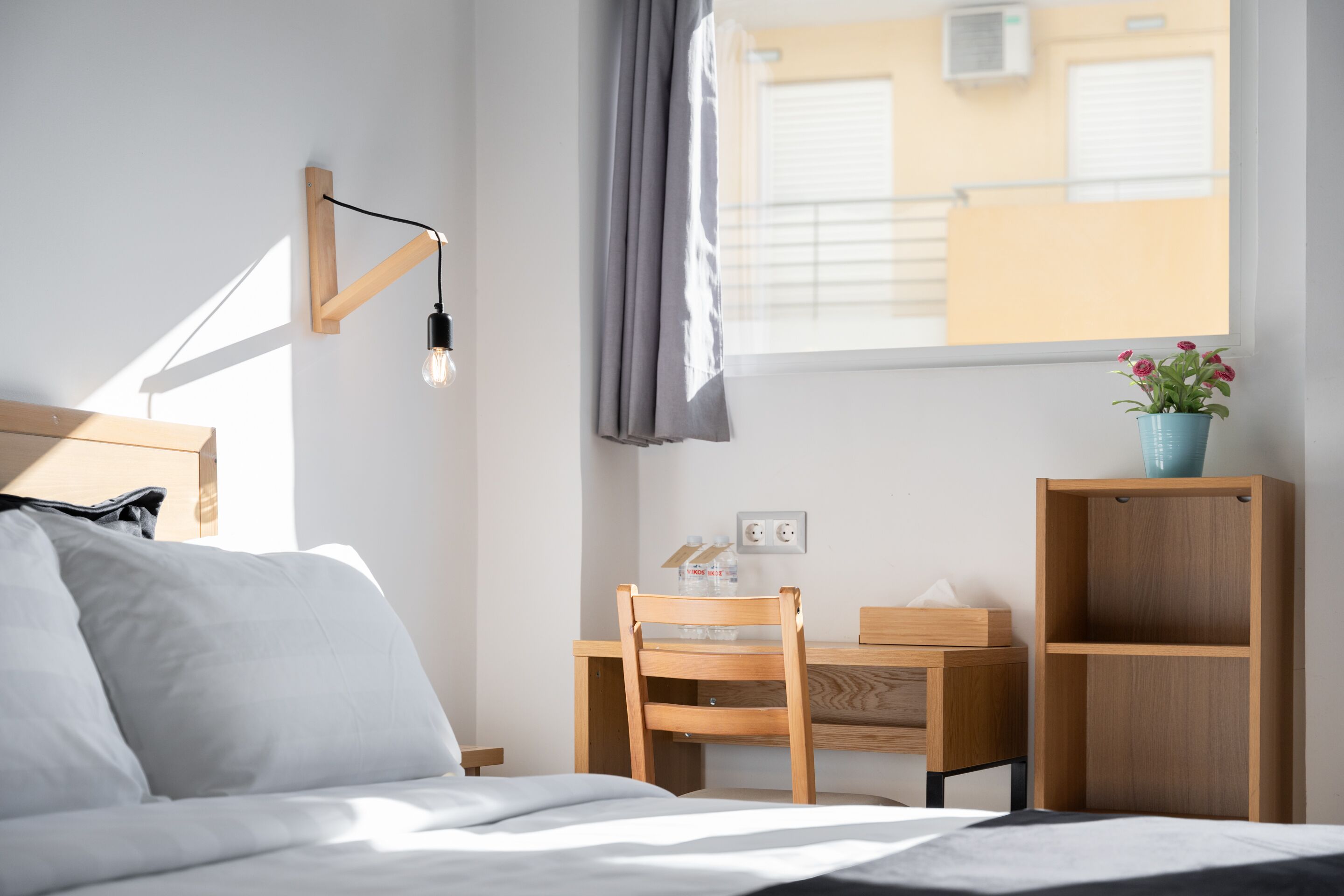 Una habitación de hotel ordenada con cama, escritorio y silla. Hay una ventana con cortinas, una lámpara de pared y una pequeña planta en el escritorio.