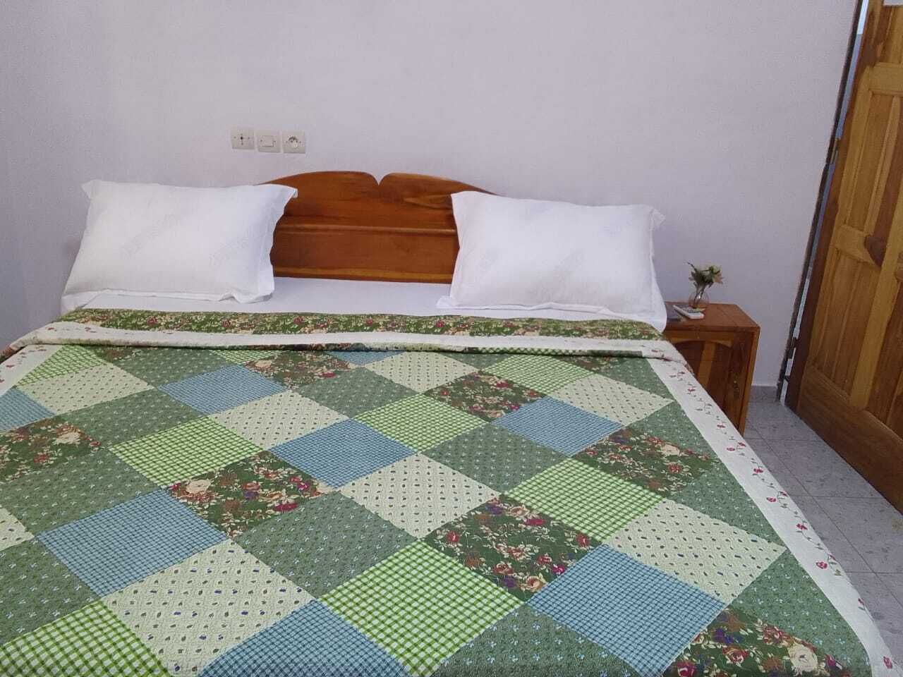 A bed with a quilt, pillows, a wooden headboard, a nightstand with a small plant, and a door.