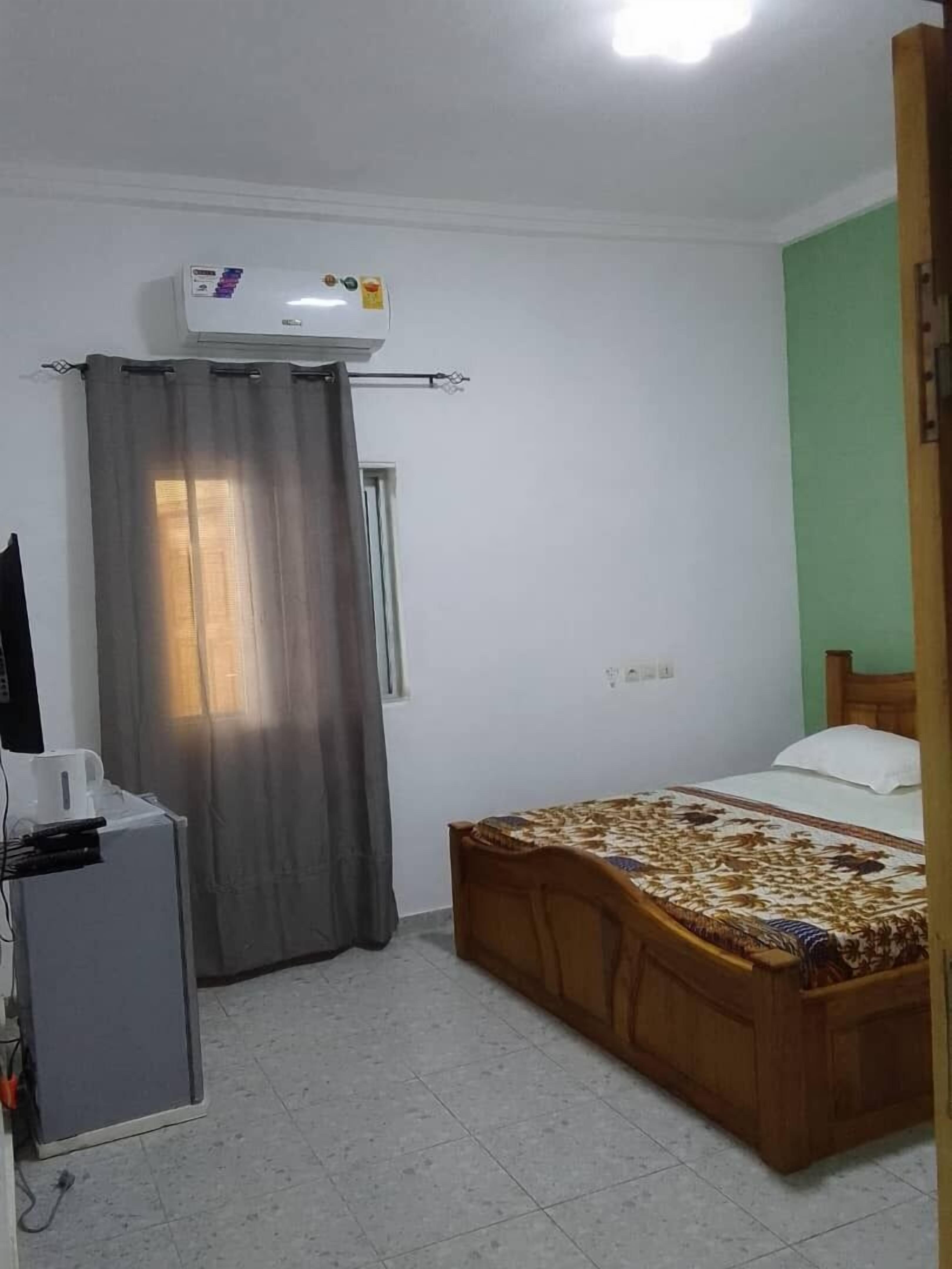 Een kleine, schone kamer met een bed, een bureau met een computer en een raam met gordijnen.