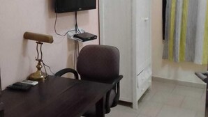 Una pequeña sala de oficina con un escritorio, una silla, un televisor montado en la pared, un armario y un perchero con toallas a rayas.