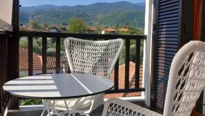 Ein Balkon mit einem weißen Korbmöbel-Tisch und -Stühlen, Blick auf grüne Hügel und eine Holztür mit Fensterläden.