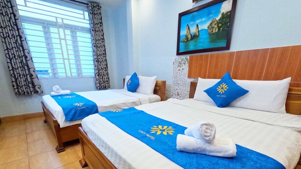 A hotel room with two beds, each with a blue bedspread and a towel. There is a painting on the wall and a window with curtains. - Cozy Hotel HCM (Ho Chi Minh City)