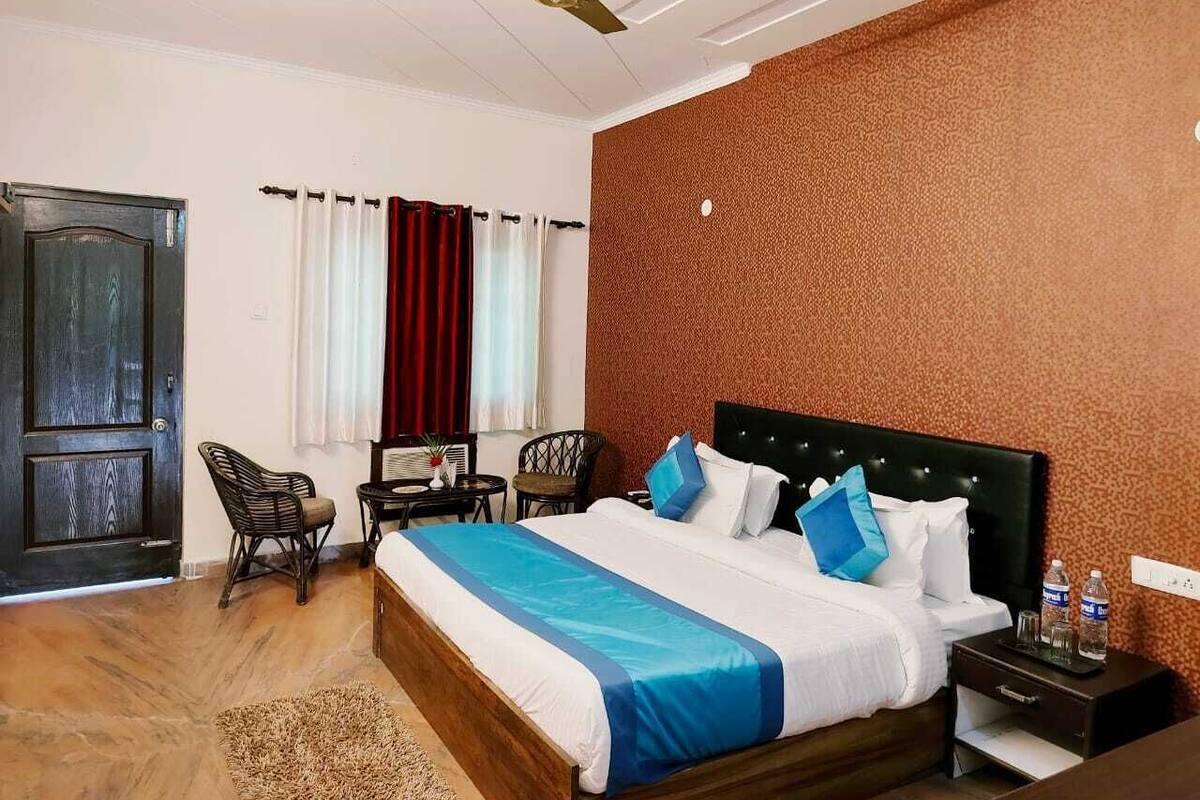 A hotel room with a large bed, a ceiling fan, a wooden floor, a red curtain, a small table with a chair, and a wall with a textured pattern.