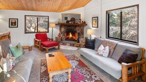 Un salon chaleureux avec une cheminée en pierre, des meubles en bois et de grandes fenêtres donnant sur la forêt.