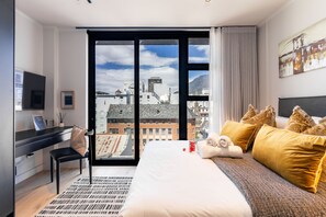 Moderni hotellihuone, jossa on suuri sänky, työpöytä ja näkymä kaupungin ylle lattiasta kattoon ulottuvien ikkunoiden kautta.