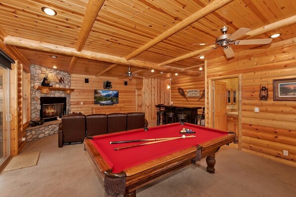 A wooden interior with a pool table, seating area, fireplace, and a ceiling fan.