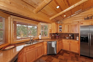A modern kitchen with wooden cabinets, stainless steel appliances, and a large window providing a view of the outdoors. - Bear Pause By Destination Big Bear: Walking Distance to the Lake! Castle Glen Estates Log Cabin Home! PET FRIENDLY! Fully Fenced! Pool Table! HOT TUB! Boarders Eagle Preserve! (Big Bear Lake)