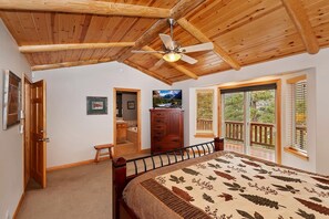 A bedroom with a wooden ceiling, a bed with a floral bedspread, a dresser, a television, and a balcony with a view of trees.