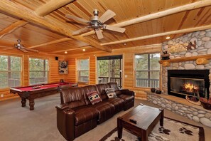 L’intérieur d’une cabane en rondins, avec une table de billard, un canapé en cuir, une cheminée et de grandes fenêtres.