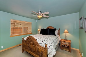 A bedroom with a bed, a ceiling fan, a bedside table with a lamp, and a window with blinds. - Bear Pause By Destination Big Bear: Walking Distance to the Lake! Castle Glen Estates Log Cabin Home! PET FRIENDLY! Fully Fenced! Pool Table! HOT TUB! Boarders Eagle Preserve! (Big Bear Lake)