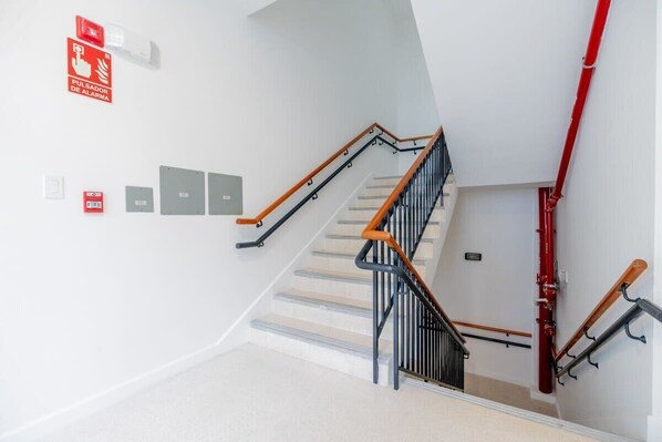 Un escalier avec des rampes en métal et des mains courantes en bois, un dispositif d’alarme incendie et un enrouleur de tuyau d’incendie rouge fixé au mur.