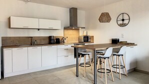 A modern kitchen with white cabinets, a wooden countertop, a built-in microwave, and a coffee maker. There are two bar stools with metal legs. - Appartement T2 Neuf, les Terrasses de Ciaccone Proche mer et Montagne (Lecci)