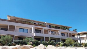 A multi-story building with balconies, large rocks in the foreground, and a clear blue sky. - Appartement T2 Neuf, les Terrasses de Ciaccone Proche mer et Montagne (Lecci)